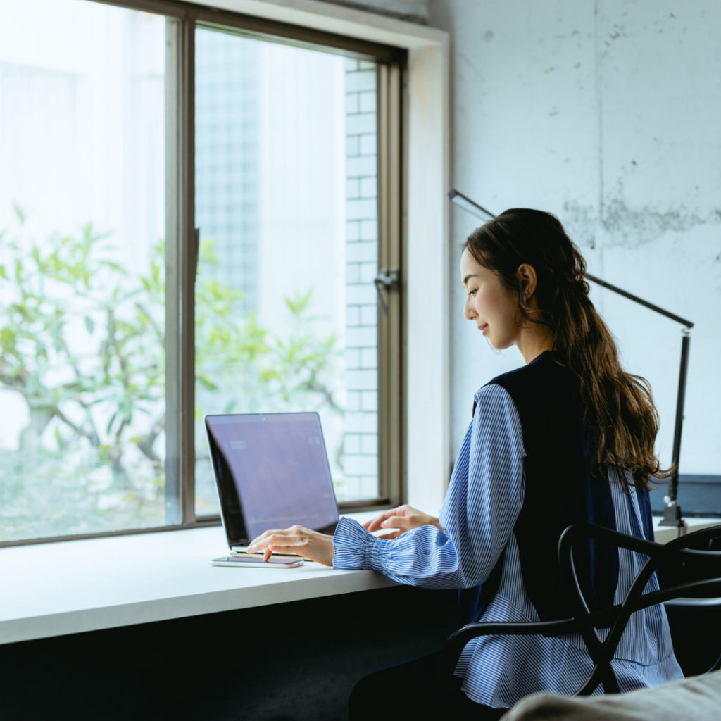A lady working on her laptop at her desk faced towards a huge window.