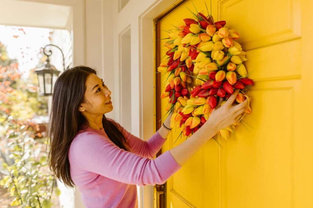 a woman hangs a pretty floral wreath on her front door in preparation to list her home